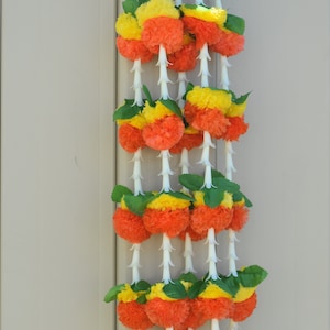 May include: A garland of orange and yellow artificial flowers with green leaves. The flowers are arranged in a vertical line and are strung together with white string.