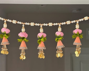 May include: A decorative garland featuring pink fabric flowers, green leaves, and peach-colored cones. The garland is strung with white beads, gold accents, and dangling gold coins. The floral arrangement is suitable for festive occasions.