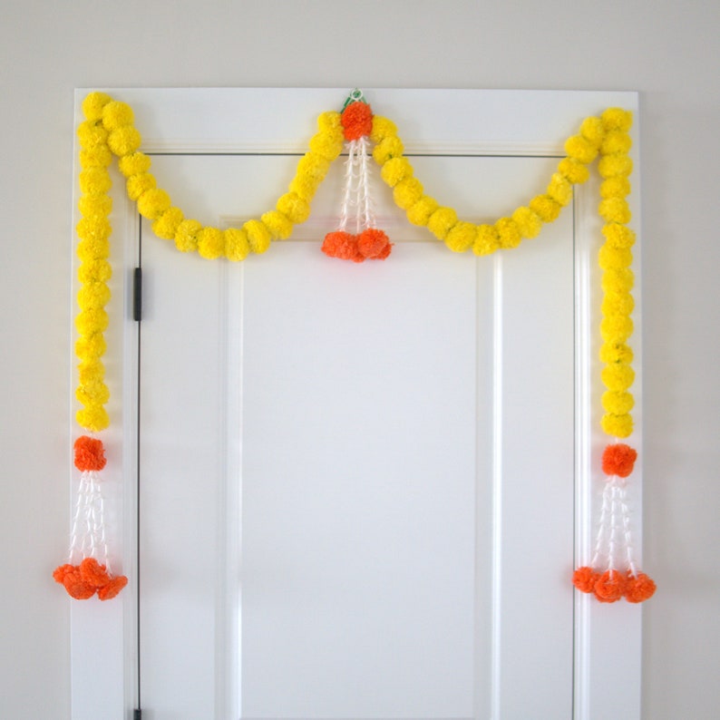 K&ouml;nnte beinhalten: Eine gelbe Ringelblumenkette mit orangefarbenen Bommeln h&auml;ngt &uuml;ber einer wei&szlig;en T&uuml;r. Die Kette ist mit wei&szlig;em Garn zusammengef&uuml;gt und hat ein gr&uuml;nes Blatt oben.