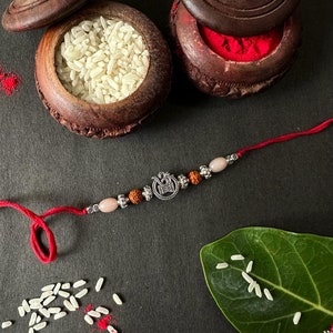 May include: A red thread bracelet featuring a central silver-toned Om symbol charm, flanked by two small brown textured beads, two smooth pale pink oval beads, and decorative silver-toned metal spacers arranged symmetrically along the cord.