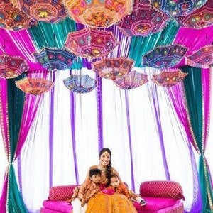 May include: A vibrant scene featuring colorful, patterned umbrellas suspended from the ceiling, creating a festive atmosphere. The umbrellas are in shades of orange, pink, and blue. A woman and two children are seated on a pink sofa.