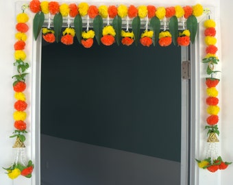 May include: A traditional Indian door decoration made with orange and yellow fabric flowers, green leaves, and white beads. The garland is hung on a white door frame.
