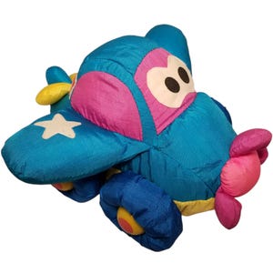 May include: A blue and pink plush airplane toy with a white star on the wing and yellow wheels. The airplane has a friendly face with black eyes and a pink mouth.
