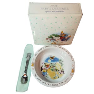 May include: A baby's keepsake spoon and bowl set. The white bowl features a nursery rhyme scene with the text "Hey Diddle Diddle! The Cat and the Fiddle." The spoon is silver-toned and rests on a light blue cloth. The box is white with a whimsical illustration.