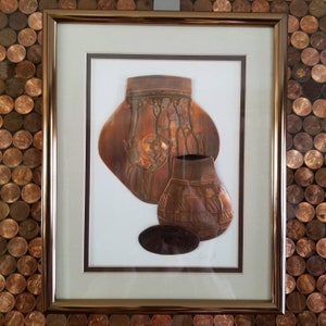 May include: A framed artwork featuring two copper-colored pottery pieces, one larger and one smaller, with intricate designs. The artwork is set against a white background and framed in a brown wood frame.