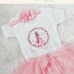 May include: A pink tulle tutu skirt and a white onesie with a bunny rabbit graphic and the text "My First Easter" and "Melody-Rae".