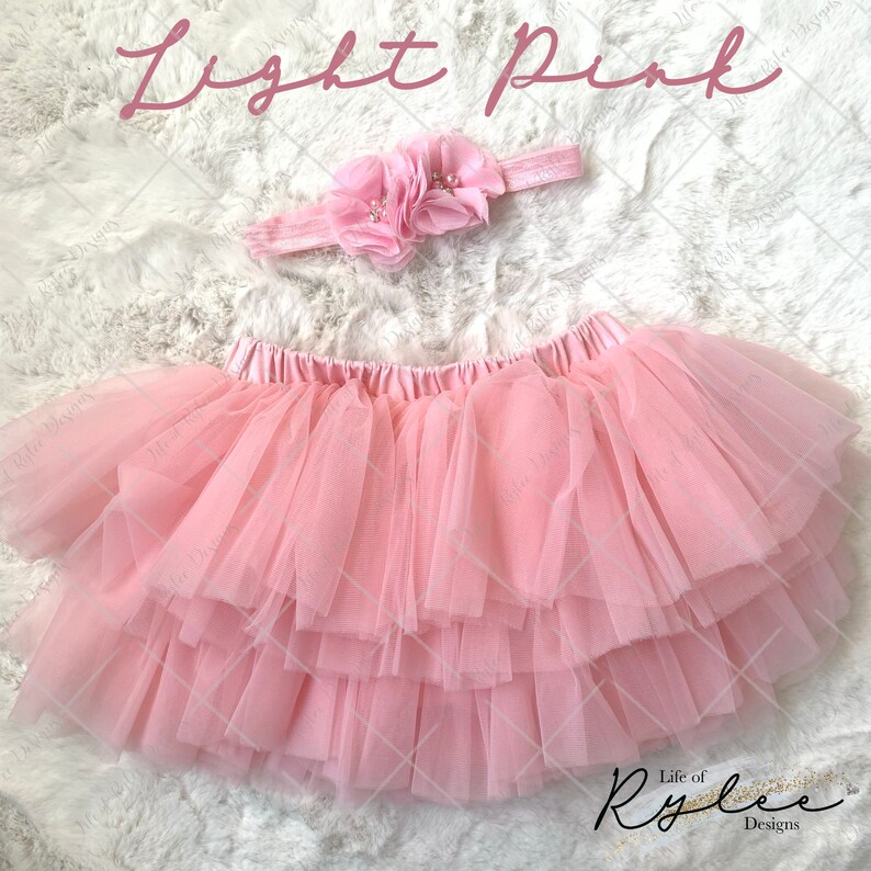 May include: Light pink tulle tutu skirt with a matching light pink flower headband. The skirt has multiple layers of tulle and a satin waistband. The headband has a cluster of three pink flowers with pearl accents.