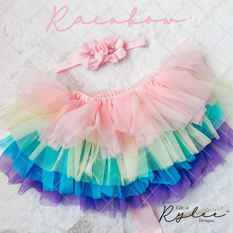 May include: Rainbow tulle tutu skirt with a pink headband and two pink flower embellishments. The headband has a pearl accent. The skirt has layers of tulle in rainbow colors, including purple, blue, green, yellow, and pink.