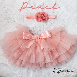 May include: Peach colored tulle tutu skirt with a matching satin bow and headband with a peach flower. The skirt is layered and has a fluffy appearance. The headband has a single flower with a pearl center.