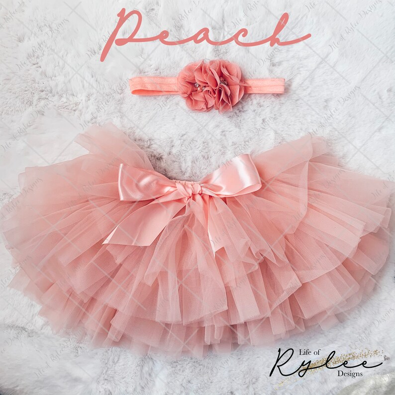 May include: Peach colored tulle tutu skirt with a matching satin bow and headband with a peach flower. The skirt is layered and has a fluffy appearance. The headband has a single flower with a pearl center.