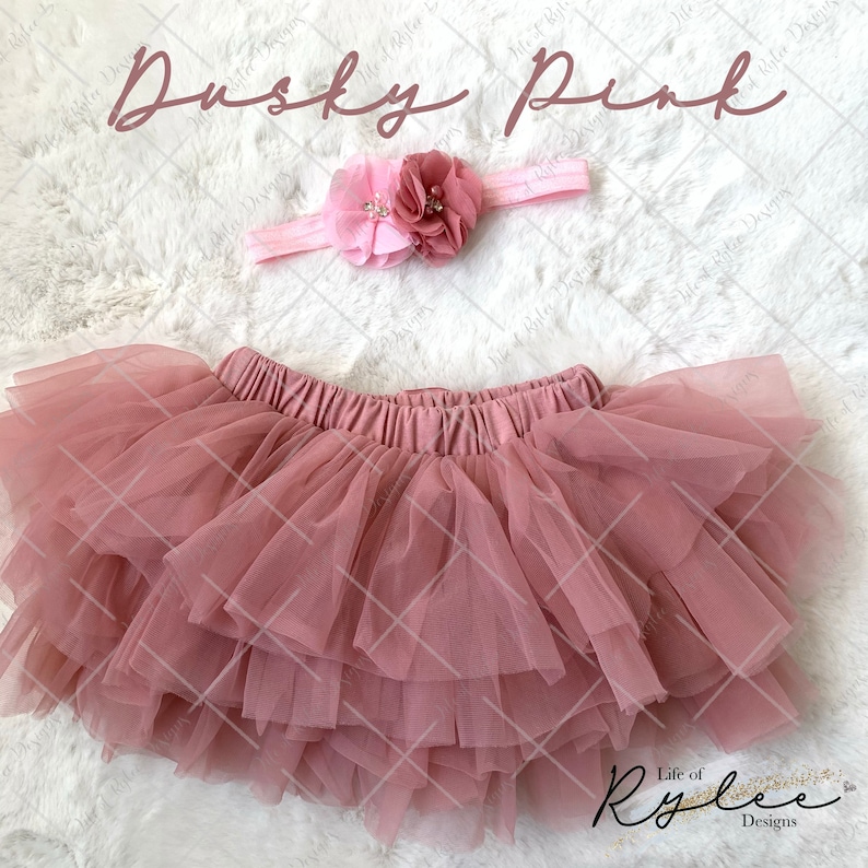 May include: A dusty pink tulle tutu skirt with a matching dusty pink headband with a pink flower accent. The headband and skirt are on a white fluffy surface. The text "Dusky Pink" is at the top of the image. The text "Life of Rylee Designs" is at the bottom of the image.