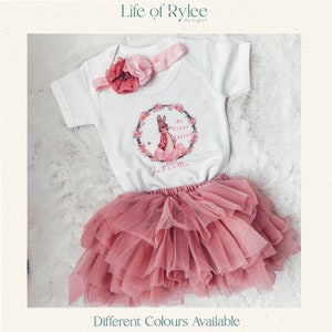 May include: A white baby onesie with a pink floral wreath and a bunny rabbit graphic. The onesie says "My First Easter" and is personalized with the name "Juliette". The onesie is paired with a pink tulle skirt.