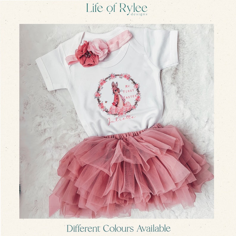 May include: A white baby onesie with a pink floral wreath and a bunny rabbit graphic. The onesie says "My First Easter" and is personalized with the name "Juliette". The onesie is paired with a pink tulle skirt.