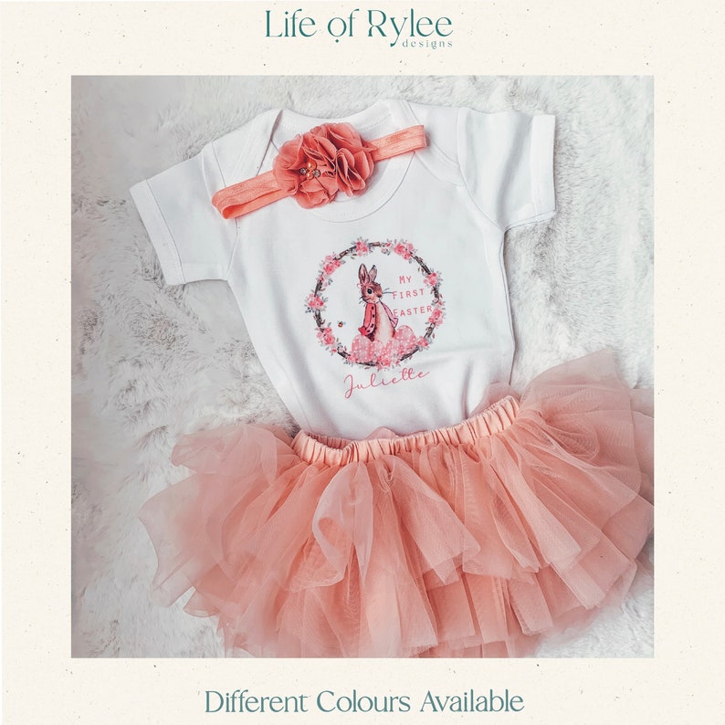 May include: A white baby onesie with a pink bunny rabbit graphic and the text "My First Easter" and "Juliette". The onesie is paired with a pink tulle skirt and a pink headband with a flower.