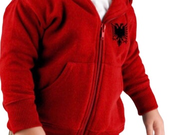May include: A red hooded zip-up sweatshirt for a child, featuring a black emblem on the chest. The sweatshirt has a full zipper, a hood, and a front pocket. The child is wearing a white shirt underneath and khaki pants.