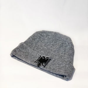 May include: A gray beanie with a black double-headed eagle emblem. The eagle is the national symbol of Albania.