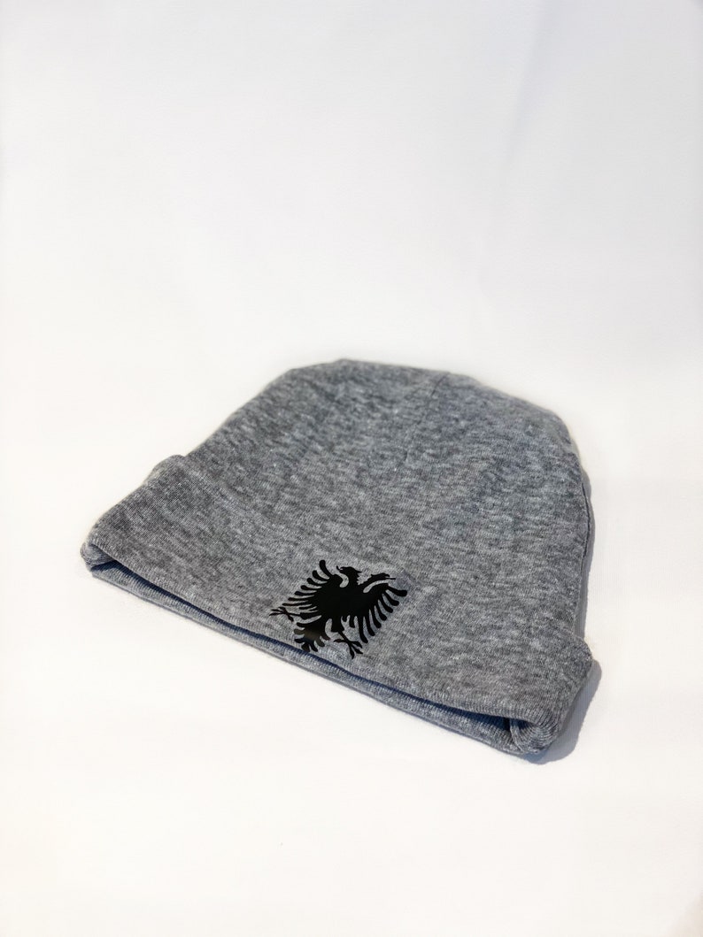 May include: A gray beanie with a black double-headed eagle emblem. The eagle is the national symbol of Albania.