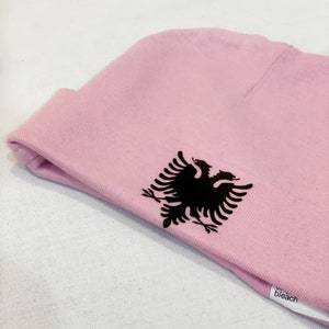 May include: A pink beanie with a black embroidered Albanian eagle on the side. The beanie has a white tag that says "bleach" on it.