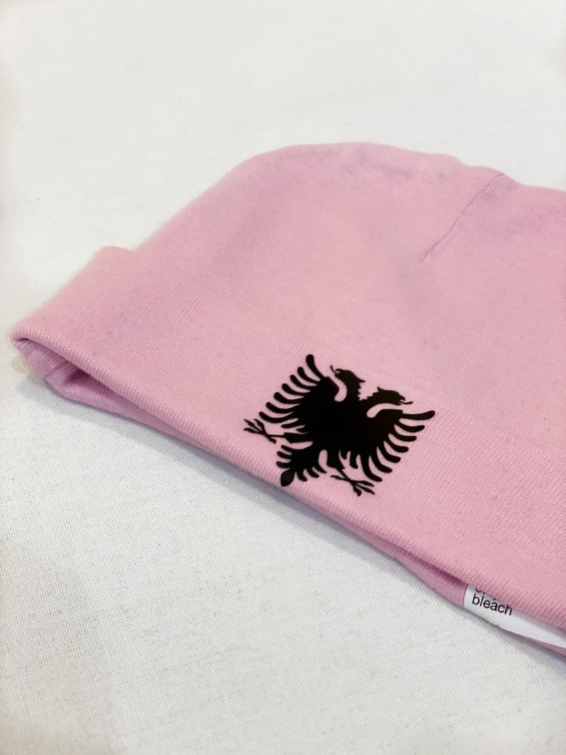 May include: A pink beanie with a black embroidered Albanian eagle on the side. The beanie has a white tag that says "bleach" on it.