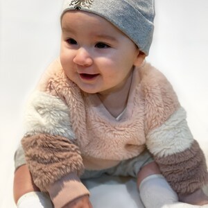 May include: A baby wearing a gray beanie with a black and silver eagle emblem, a pink and white fuzzy sweater, and white socks.