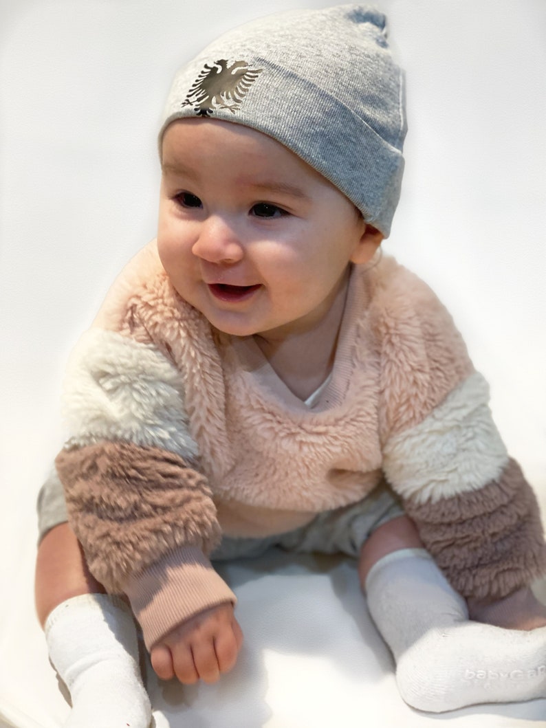 May include: A baby wearing a gray beanie with a black and silver eagle emblem, a pink and white fuzzy sweater, and white socks.