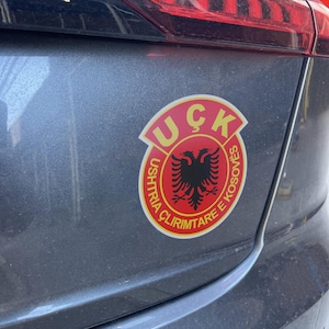 Może przedstawiać: Naklejka w kolorze czerwonym i żółtym z napisem "UCK" i "USHTRIA CLIRIMTARE E KOSOVES" w kółku wokół czarnego dwugłowego orła.