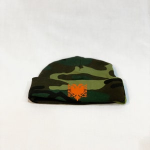 May include: A green, brown, and black camouflage beanie with an orange Albanian eagle emblem.