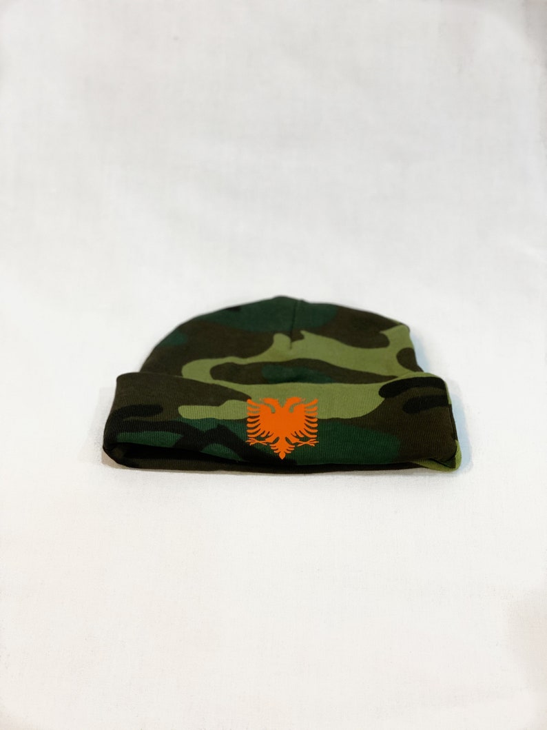 May include: A green, brown, and black camouflage beanie with an orange Albanian eagle emblem.