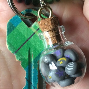 May include: A small glass bottle with a cork stopper filled with gray and purple painted stones. The bottle is attached to a key ring.