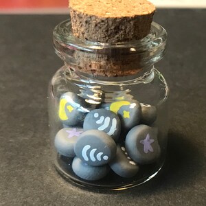 May include: A glass jar with a cork lid filled with small gray clay beads. Each bead has a different design, including stars, moons, and wavy lines.
