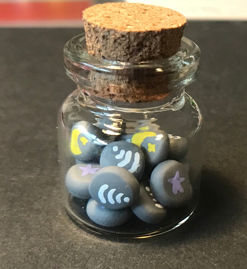 May include: A glass jar with a cork lid filled with small gray clay beads. Each bead has a different design, including stars, moons, and wavy lines.