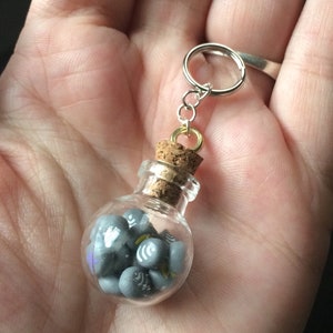 May include: A small glass bottle with a cork stopper filled with gray and white miniature charms. The bottle is attached to a silver key chain.