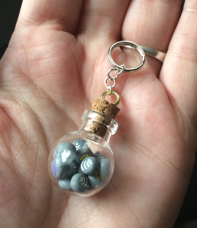 May include: A small glass bottle with a cork stopper filled with gray and white miniature charms. The bottle is attached to a silver key chain.
