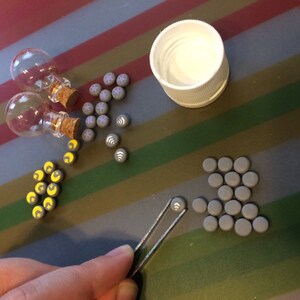 May include: Assortment of small gray clay tiles with various symbols, including moons, stars, and wifi symbols. Two small glass bottles with cork stoppers and a white lid are also included.