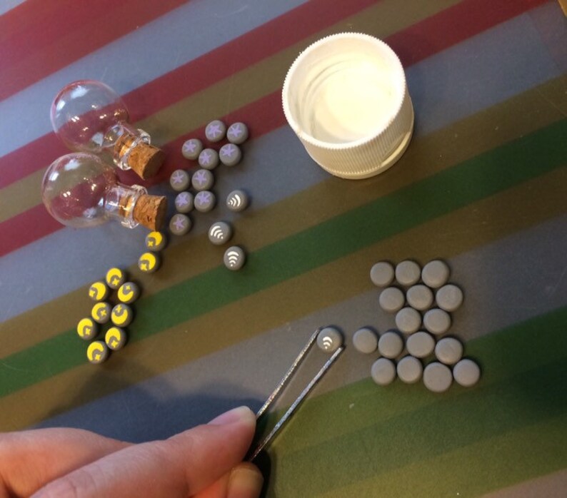 May include: Assortment of small gray clay tiles with various symbols, including moons, stars, and wifi symbols. Two small glass bottles with cork stoppers and a white lid are also included.