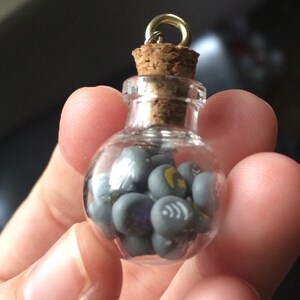 May include: A small glass bottle with a cork stopper and a gold chain. The bottle is filled with gray and silver charms with various symbols.