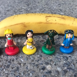 May include: Seven small figurines, each standing on a colored base, are arranged in a row. The figurines are made of clay and depict a scuba diver, a construction worker, a woman with long hair, a creature with green skin and a cloak, a woman with goggles, and a man in a suit. A yellow banana is behind the figurines.