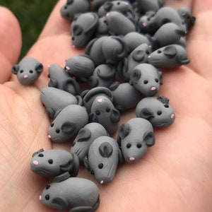 Op de afbeelding: Een verzameling kleine grijze polymer clay muizen met roze neuzen. De muizen zijn op een hand van een persoon in een stapel gerangschikt.