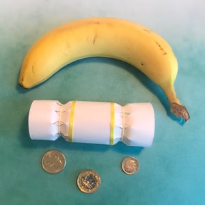 May include: A white paper Christmas cracker with yellow ribbon, a yellow banana, and three gold coins on a teal background.