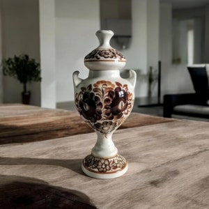 May include: A white ceramic vase with a brown floral design. The vase has a round body and a narrow neck with a lid. The vase is sitting on a wooden table.