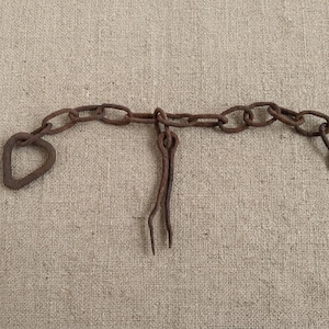 May include: A rusty brown metal chain with a heart-shaped loop on one end and a loop on the other. A metal piece with two prongs is attached to the chain. The chain is lying on a tan fabric surface.