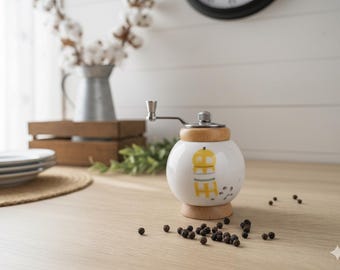 May include: A white ceramic pepper grinder with a wooden base and top. The grinder features a yellow and gray design. Black peppercorns are scattered on the light wood surface. A metal handle is attached to the top.