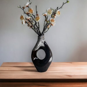 May include: A black ceramic vase with a unique, abstract shape and a hole in the side. The vase is filled with white flowers with yellow centers.