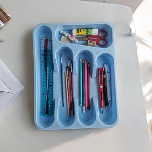 K&ouml;nnte beinhalten: Ein blauer Plastik-Besteck-Organizer mit sechs F&auml;chern. Der Organizer enth&auml;lt ein Lineal, Pinsel, Bleistifte und Stifte. Der Organizer befindet sich auf einer wei&szlig;en Oberfl&auml;che.