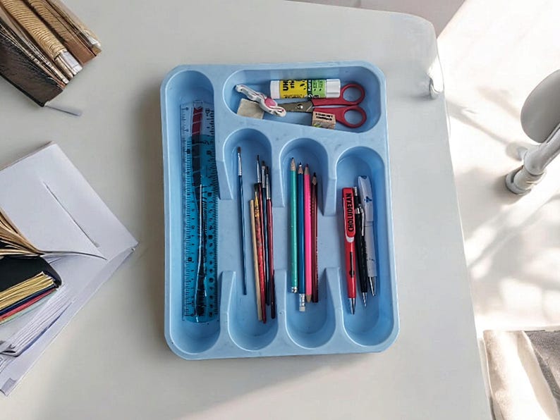 K&ouml;nnte beinhalten: Ein blauer Plastik-Besteck-Organizer mit sechs F&auml;chern. Der Organizer enth&auml;lt ein Lineal, Pinsel, Bleistifte und Stifte. Der Organizer befindet sich auf einer wei&szlig;en Oberfl&auml;che.