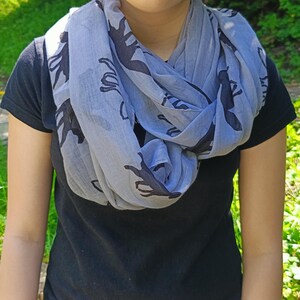 May include: A light blue infinity scarf with a repeating black silhouette pattern. The scarf is draped around the neck, creating multiple layers. The person is wearing a black t-shirt. The background is blurred with green foliage.