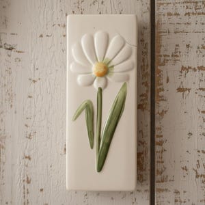 May include: A rectangular, cream-coloured ceramic wall plaque with a raised daisy design. The daisy has white petals, a yellow centre, and green leaves and stem. The plaque is mounted on a distressed, white wooden surface.