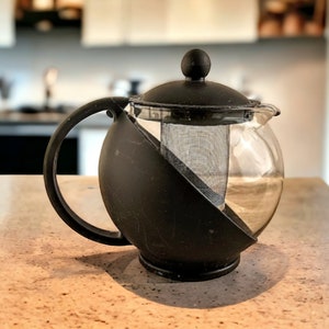 May include: A black and clear glass teapot with a built-in strainer. The teapot has a round body and a black handle.