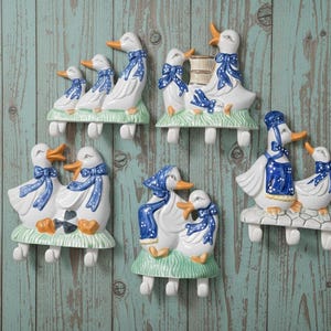 Può includere: Una collezione di statuine di anatra in ceramica, ognuna con un design unico. Le anatre sono bianche con dettagli blu, tra cui fiocchi e copricapi. Montate su basi verdi con ganci, adatte per essere appese.