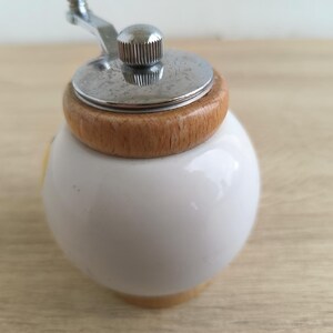 A small, round vintage-style pepper mill with a white porcelain body and a light wooden top. The mill features a subtle yellow geometric graphic on its side and has an exposed metal grinding mechanism at the top. The image is a close-up, showcasing the mill's design and internal structure, resting on a rustic farmhouse wooden table.